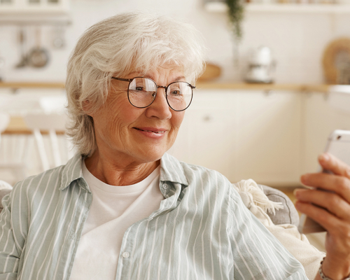 Happy joyful female pensioner in round eyeglasses surfing internet on cell phone, looking at mobile's screen with broad smile, booking plane tickets, planning trip or scrolling pics via social network